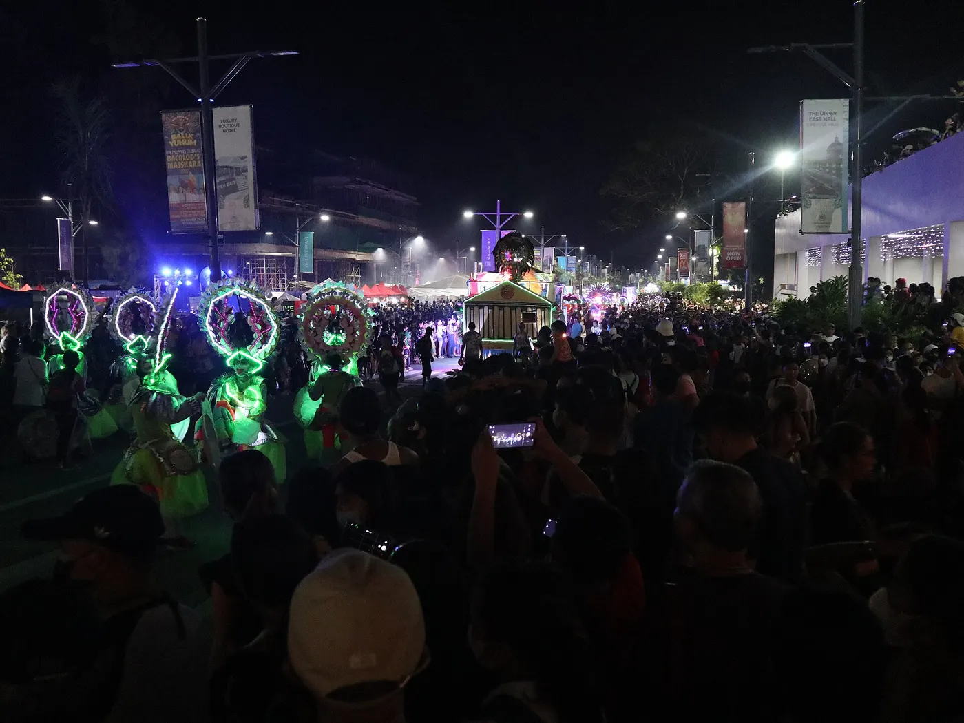 Electric MassKara dancers in glowing LED costumes and headdresses parading at night past a dense crowd at The Upper East in Bacolod